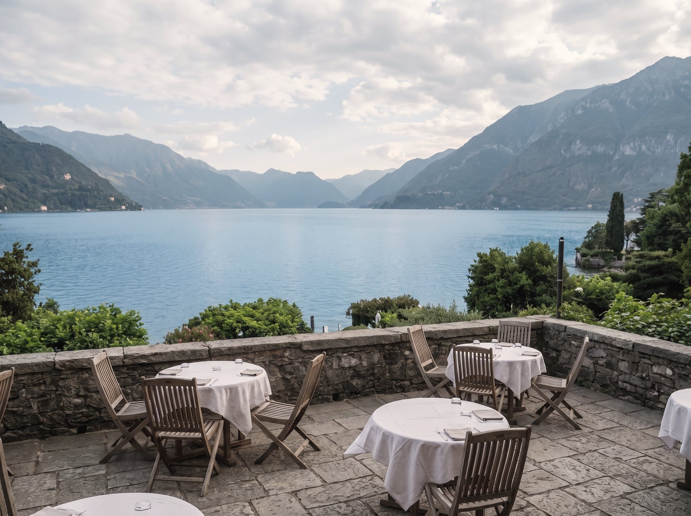 La terraza de un restaurante a orillas de un lago, con mesas vestidas de lino blanco y, al fondo, el agua, las montañas y un cielo abierto